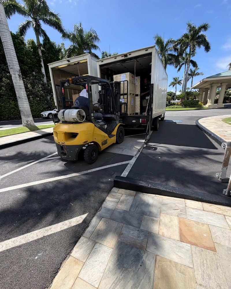 A forklift unloading a box truck in a resort driveway lined with palm trees
